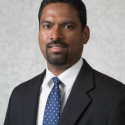 A man looks into a  camera. He is wearing a black suit, white shirt and blue and white polka dot tie.